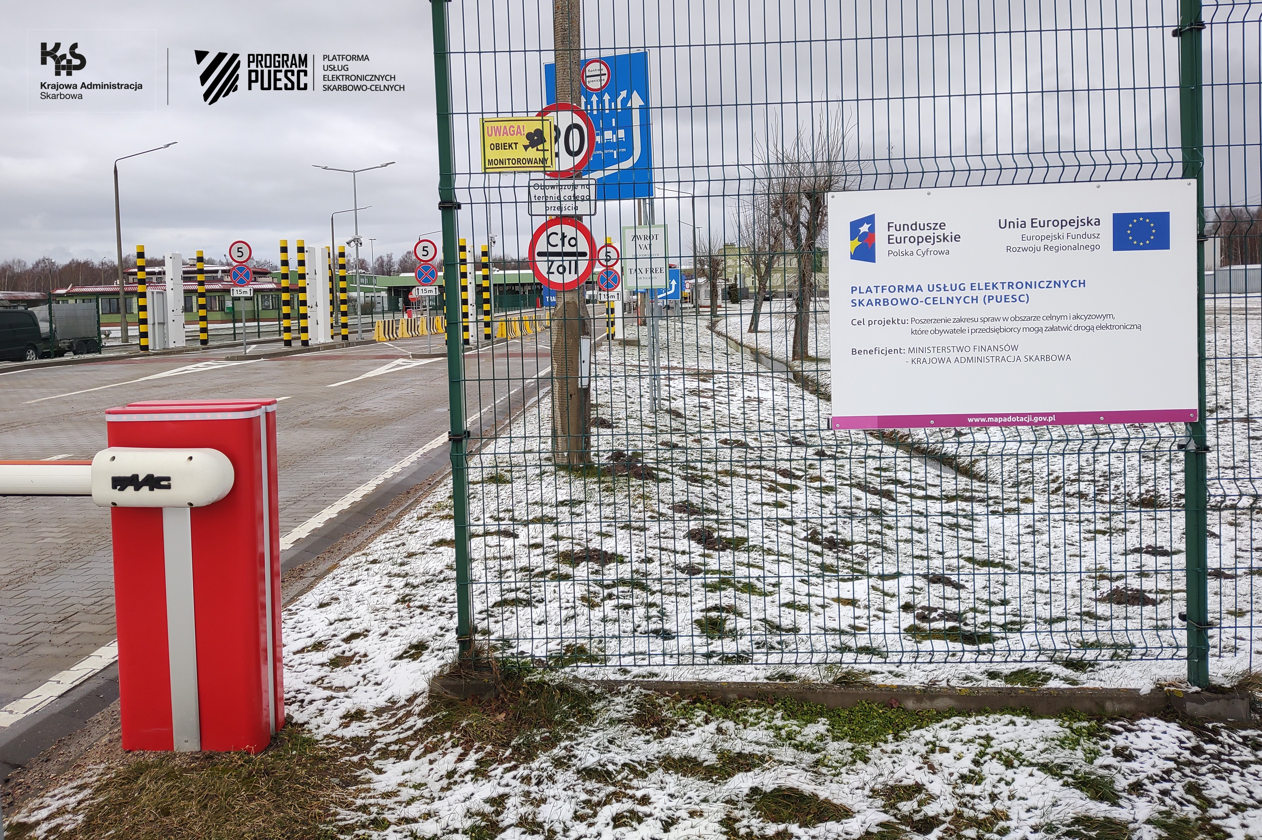 On the right side you can see a metal fence with a board informing about the PUESC Project, on which logos are placed: National Revenue Administration, European Funds, Digital Poland, Tax and Customs Information System, Ministry of Finance, European Union European Regional Development Fund. On the left, at the bottom, there is a barrier, in the background you can see yellow and black pillars and road lanes.