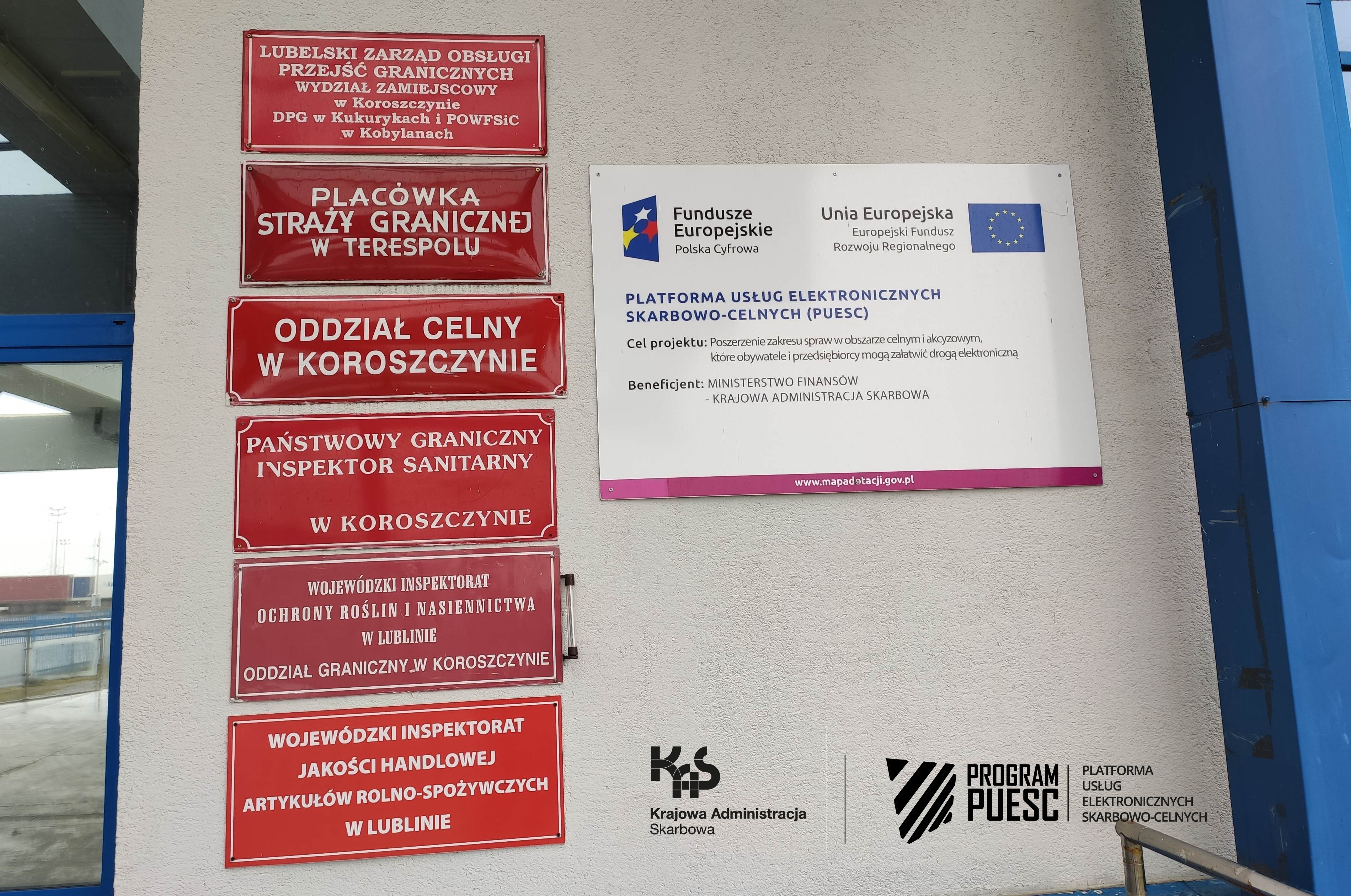In the middle of the page you can see a wall with red signs informing about the institutions located in the building. On one of the boards there is information about the PUESC Project and logos: National Revenue Administration, European Funds Digital Poland, Tax and Customs Information System, Ministry of Finance, European Union European Regional Development Fund.