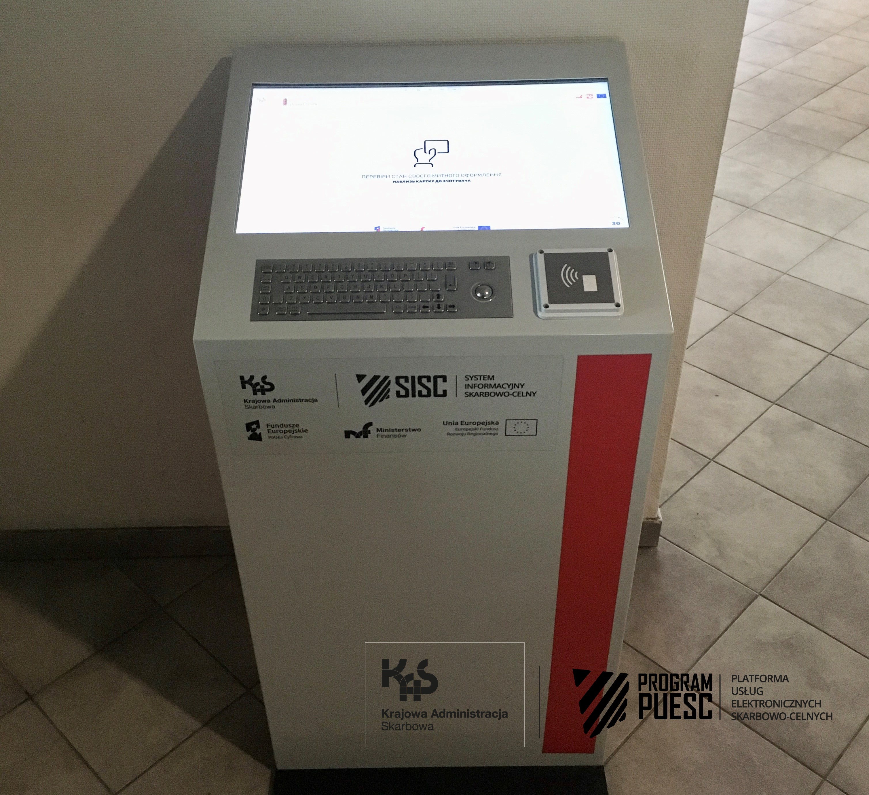 The first kind of kiosk inside the customs branch office. A device in the shape of a cuboid with a screen, a keyboard and a card reader, on which logos are placed: National Revenue Administration, European Funds Digital Poland, Tax and Customs Information System, Ministry of Finance, European Union European Regional Development Fund.